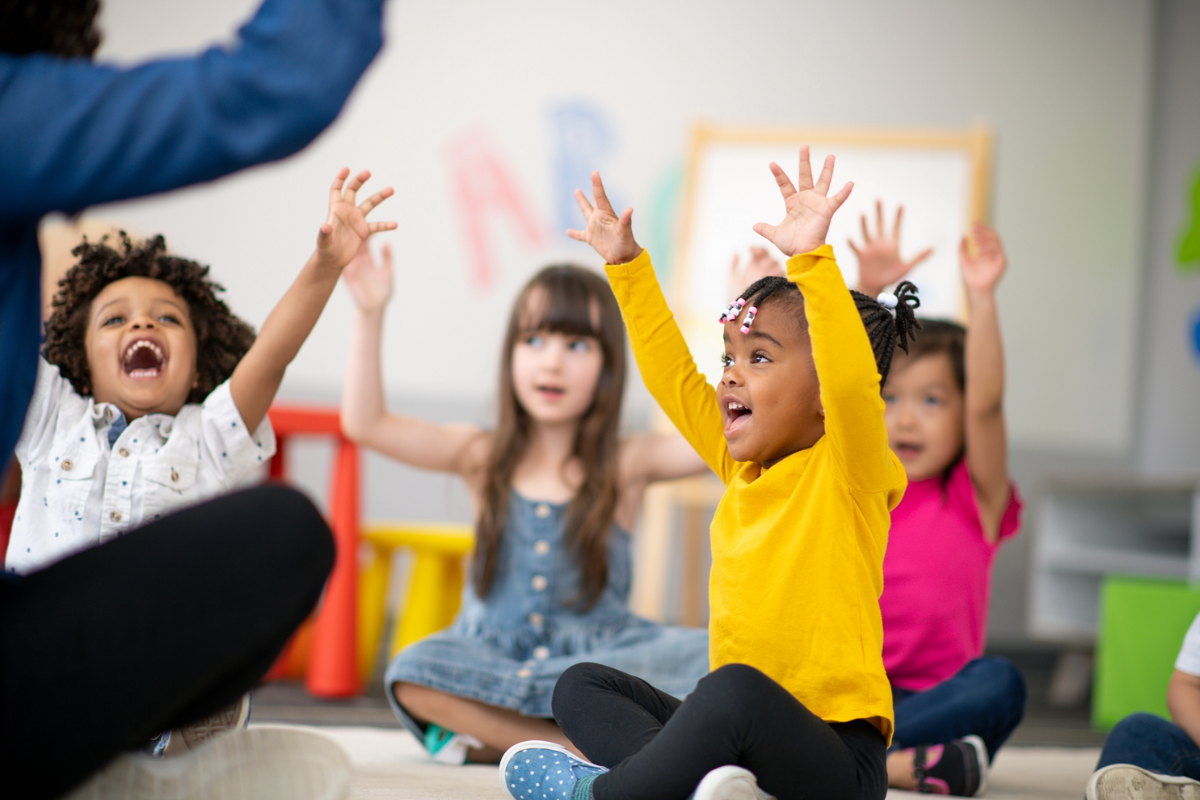 Teacher with a group of children smiling and visibly excited with both hands raised in the air.
