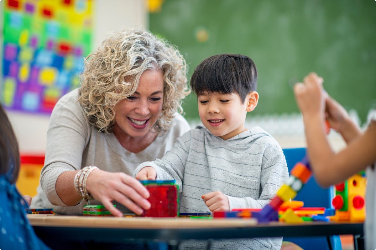 An educator and a young student are smiling while playing with blocks at a table.